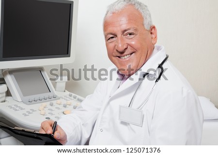 Portrait of happy senior male radiologist writing on clipboard with ultrasound machine in background
