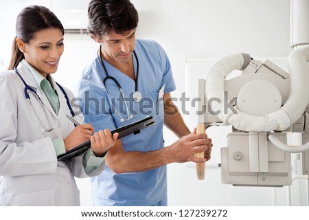 Female radiologist writing down notes while male technician setting up machine to take x-ray
