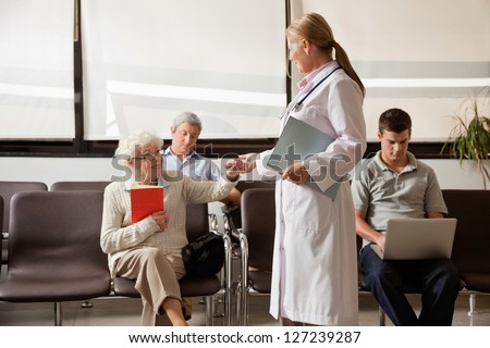 Mature female doctor holding hand of senior woman while people sitting in hospital lobby