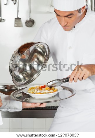 Young chef checking pasta dish with tong in commercial kitchen