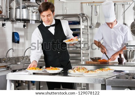 Young waiter placing dishes in tray with chef working in commercial kitchen