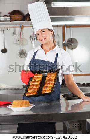 Portrait of happy female chef holding tray of baked breads in industrial kitchen