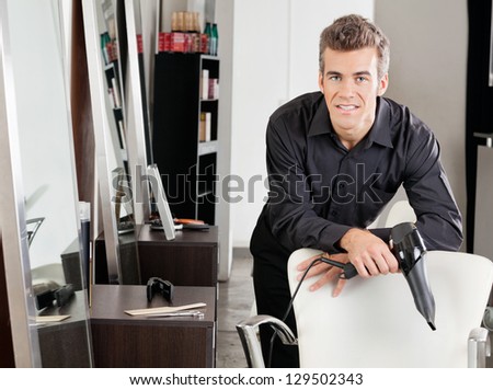 Portrait of mature male hairstylist with hairdryer leaning on chair at salon