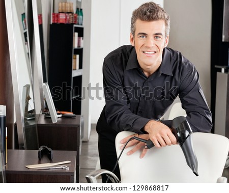 Portrait of happy hairstylist with blower leaning on chair at salon