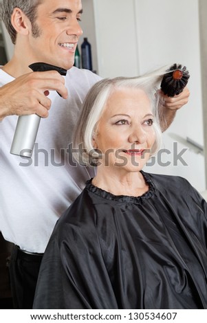 Male hairstylist with spray and hairbrush setting up woman\'s hair at beauty salon
