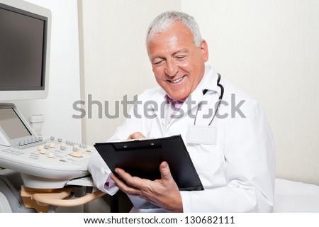 Happy senior male radiologist holding clipboard while sitting in clinic