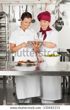 Two female chefs looking for recipe on a digital tablet while cooking at kitchen