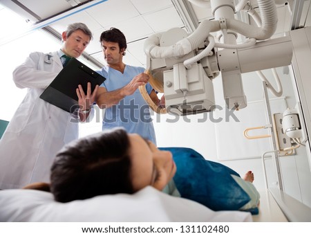 Radiologist with male technician looking at clipboard while setting up the machine to x-ray female patient