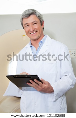 Portrait of happy mature male doctor holding clipboard while standing next to CT scan machine