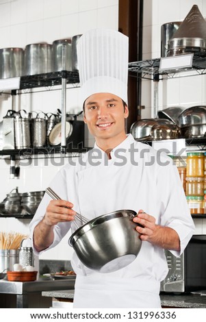Portrait of young male chef with wire whisk and mixing bowl standing at commercial kitchen