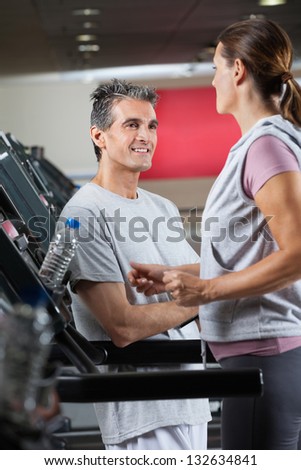 Happy mature instructor looking at female client exercising on treadmill in health center
