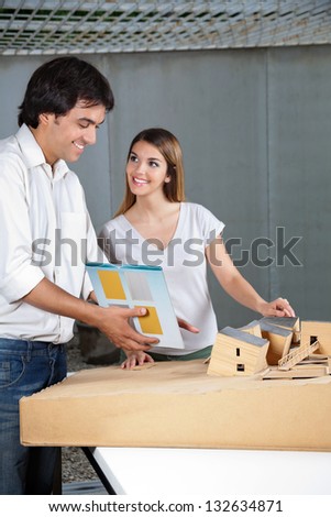 Young male architect looking at model house while female colleague smiling at him
