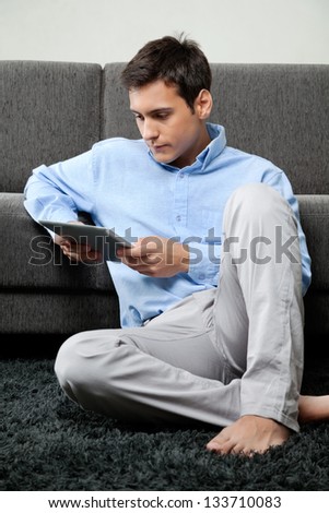 Young man in formal wear looking down at digital tablet while sitting on rug at home
