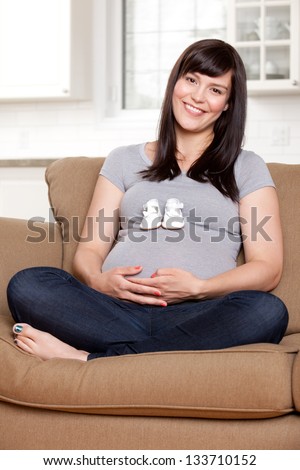 Portrait of expecting mother sitting on sofa with baby shoes on belly