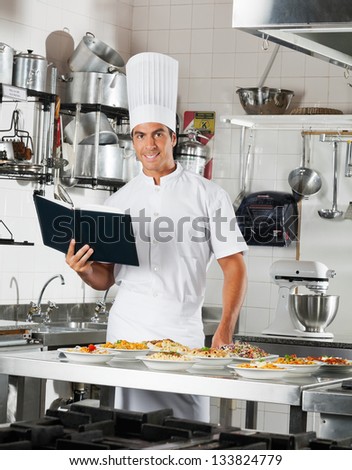 Portrait of young male chef with book standing by commercial kitchen counter