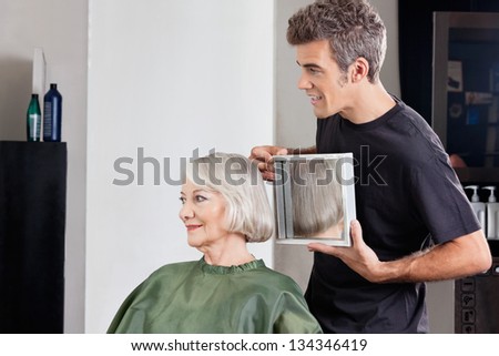 Mature male hairstylist showing finished haircut to senior female client at beauty parlor