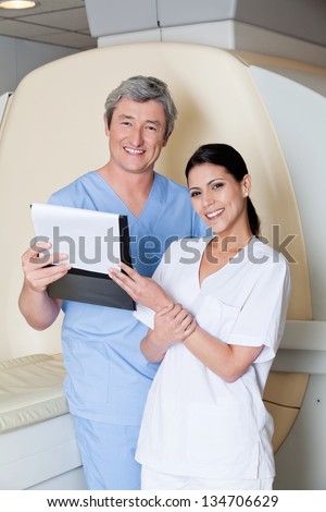 Portrait of happy multiethnic radiologic technicians holding clipboard while standing by MRI scan machine
