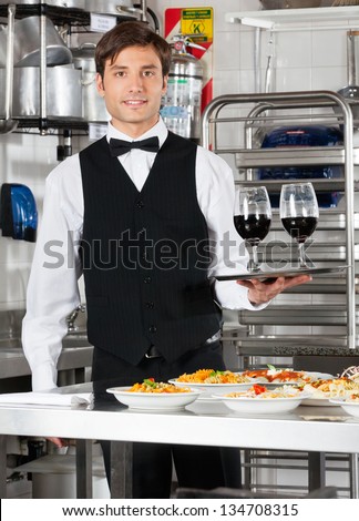 Portrait of young waiter holding wineglasses in tray with pasta dishes on commercial kitchen counter
