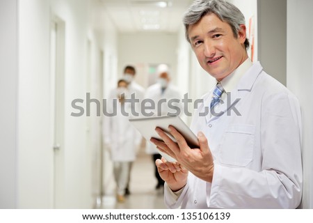 Portrait of mature male doctor holding digital tablet with colleagues walking in background