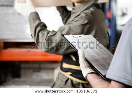 Midsection of foreman loading cardboard box with supervisor holding clipboard at warehouse