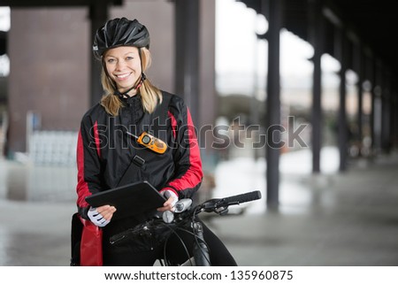Portrait of young female cyclist in protective gear with courier bag using digital tablet
