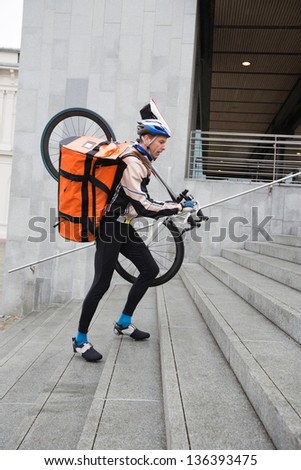 Side view of male cyclist with courier bag and bicycle on shoulder walking up steps