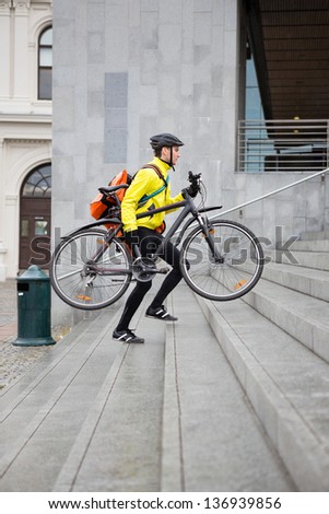 Side view of courier delivery man in protective gear with bicycle and backpack walking up steps