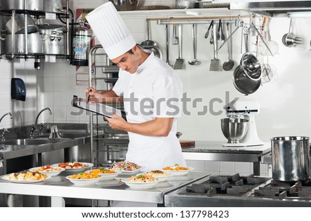 Young Male Chef With Clipboard Going Through Cooking Checklist At Commercial Kitchen Counter