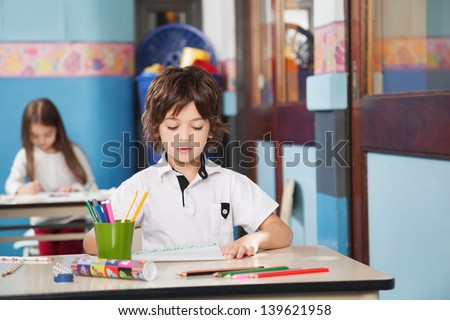 Little boy with color pencils and drawing paper while girl in background at kindergarten
