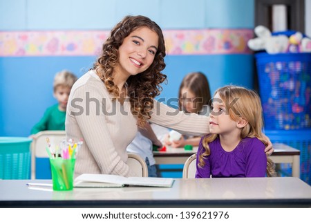 Portrait of beautiful kindergarten teacher with little girl sitting at desk in classroom