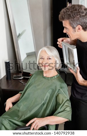 Portrait of happy senior woman with hair stylist holding mirror in salon