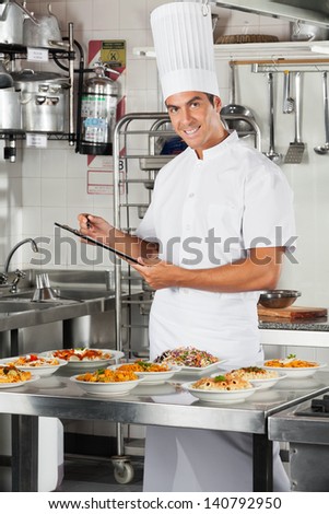 Portrait of happy male chef holding clipboard with pasta dishes on commercial kitchen counter