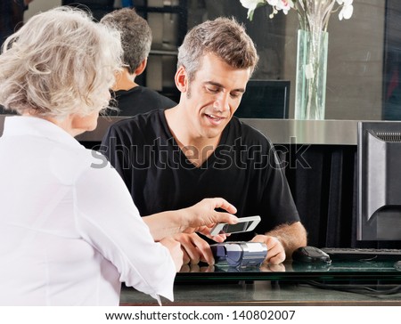 Hairdresser with female client paying with mobilephone over electronic reader at counter