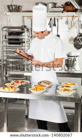 Male chef with clipboard checking pasta dishes in commercial kitchen