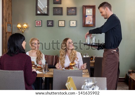Young waiter with tray of glasses while female customers having meal in restaurant