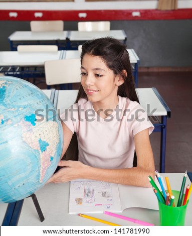 Cute little schoolgirl searching places on globe at desk in classroom