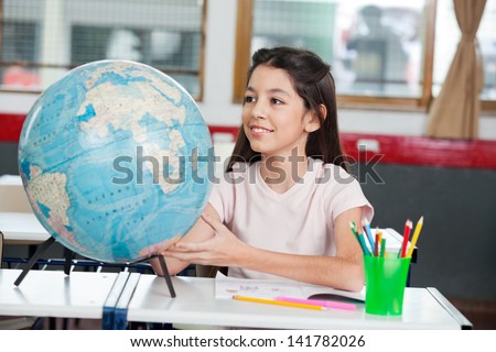 Cute little schoolgirl searching places on globe at desk in classroom