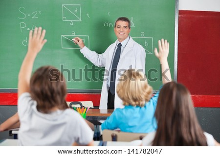 Mature male teacher teaching while students raising hands in classroom