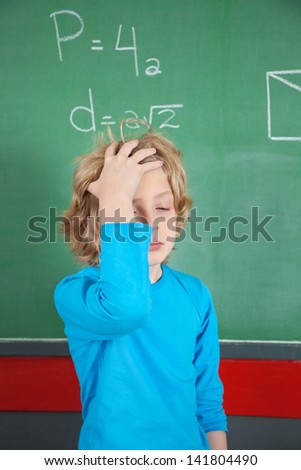 Sad little schoolboy standing with hand in hair by board in classroom