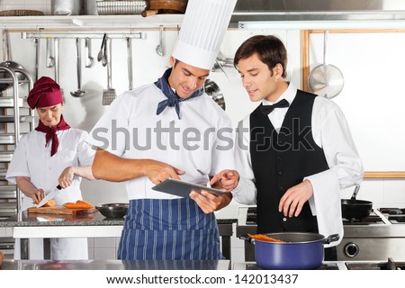 Chef And Waiter Using Digital Tablet With Female Chef Working In Restaurant Kitchen