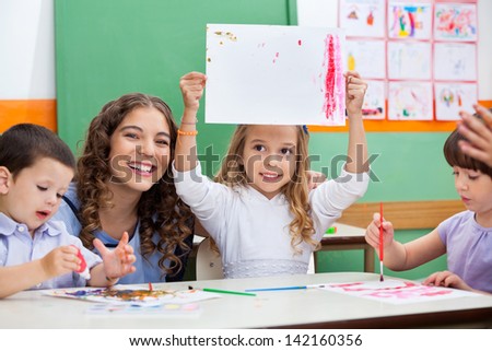 Portrait of young teacher with girl showing drawing while students painting at desk