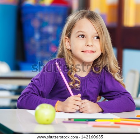 Thoughtful little girl with sketch pen and paper sitting at desk in classroom