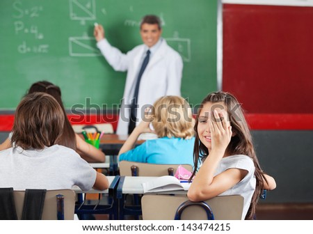 Portrait of little girl covering one eye while sitting at desk with teacher teaching in classroom