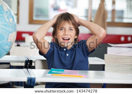 Portrait of shocked little boy sitting with globe and books at desk in classroom