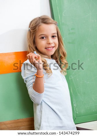 Portrait of cute girl with chalk standing by chalkboard in class