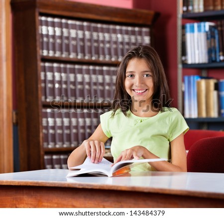 Portrait of happy cute schoolgirl with book sitting at table in library
