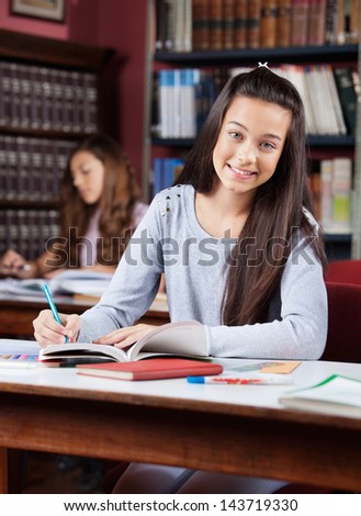 Portrait of beautiful teenage schoolgirl smiling while writing in book at table in library