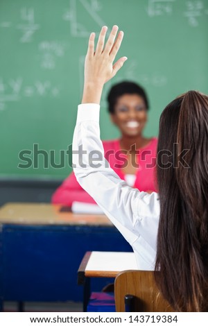 Rear view of teenage schoolgirl raising hand at desk with teacher in background