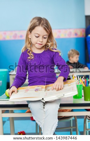 Little girl reading book while sitting on desk with friend in background at kindergarten
