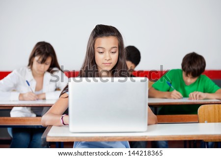 Teenage schoolgirl using laptop at desk with classmates in background